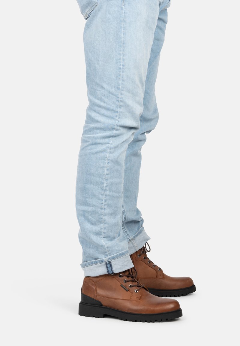 Persona che indossa jeans azzurri e stivali in pelle marrone con lacci, in piedi su uno sfondo bianco.