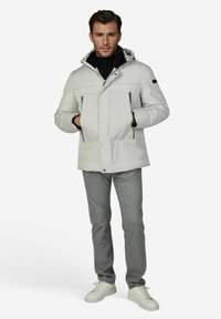 Lichtgrijze geïsoleerde jas met capuchon, voorzien van steekzakken aan de voorkant, een ritssluiting en een zwarte kraag. Gecombineerd met grijze broek en witte sneakers.