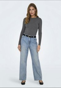 Un top gri cu mâneci lungi, blugi denim ușor albastri, lejeri, o curea neagră cu cataramă circulară și pantofi plano negri. Fundal alb simplu.