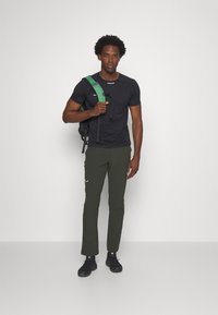 Schwarzes Sportshirt, grüner Rucksack, grüne Hose mit weißem Logo und schwarze Schuhe. Das Model steht vor einem schlichten weißen Hintergrund.