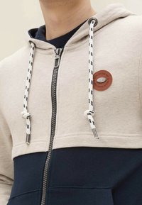 Uomo che indossa una felpa con zip bicolore beige e navy, con lacci bianchi e una toppa rotonda marrone sul petto.