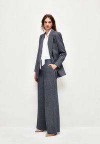 Grauer Hahnentrittmuster-Blazer mit Doppelreiher-Verschluss, kombiniert mit grauen, weit geschnittenen Hosen; darunter eine weiße Bluse; beige High Heels.