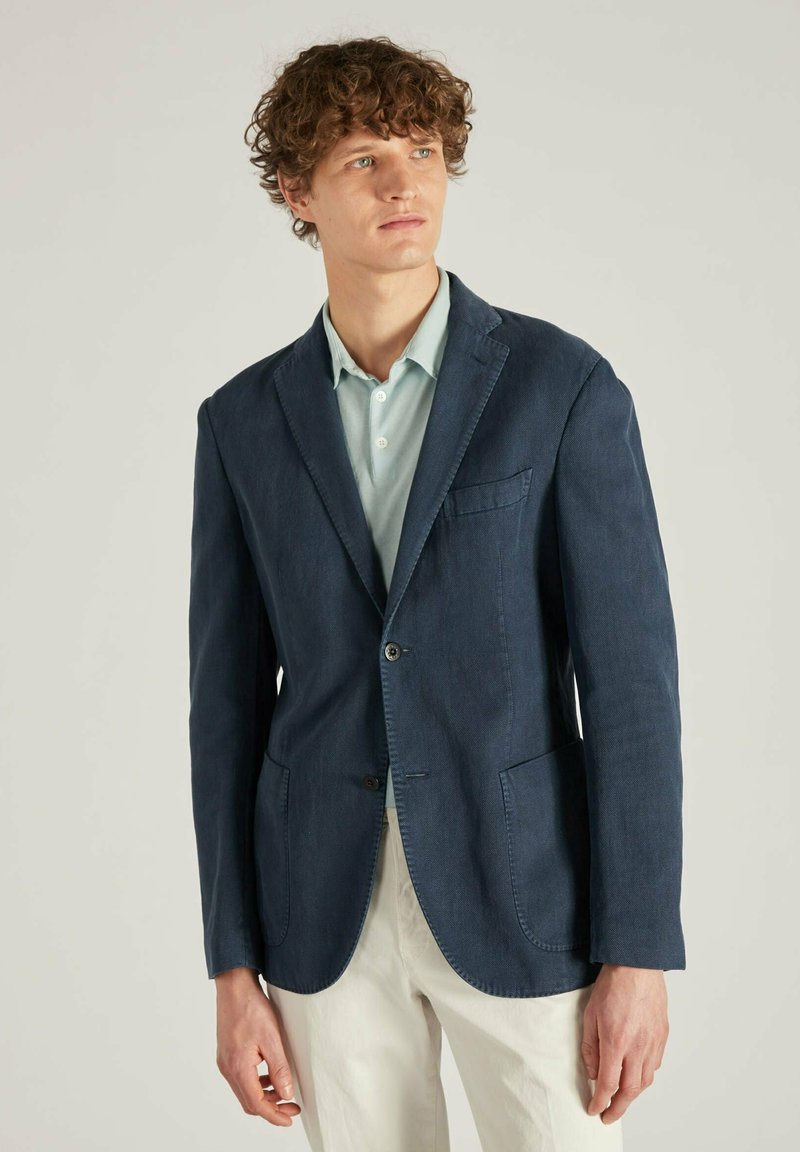 Marineblauwe op maat gemaakte blazer, lichtgewicht stof, enkele knoopsluiting, gelapte revers, twee voorkruiken, gecombineerd met een lichtblauw overhemd.