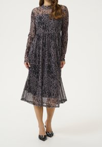 Robe à manches longues en tissu noir transparent avec un motif léopard gris. Jupe évasée, col rond et détail de taille froncée. Chaussures noires à talons pointus.