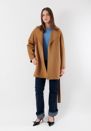 Cappotto cammello con rever, maniche lunghe e apertura frontale. Abbinato a un maglione blu, jeans in denim scuri con orli risvoltati e décolleté nere.