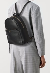 Černý kožený batoh s kulatým tvarem, viditelným zlatým zipem, přední kapsou a nastavitelnými popruhy. Nesený přes kostkovanou košili.