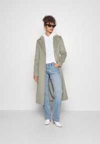 Donna che indossa un lungo cappotto grigio sopra una felpa bianca, jeans azzurri chiari, stivali bianchi con tacco e occhiali da sole tartarugati, in piedi contro uno sfondo bianco.