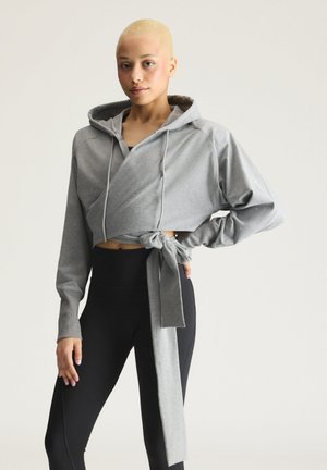 Jeune personne aux cheveux blonds courts portant un sweat à capuche gris court noué à la taille et des leggings noirs, se tenant devant un fond uni.