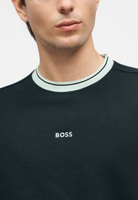 Gros plan d'une personne portant un chemisier noir avec un col rayé vert clair et blanc, et le mot « BOSS » brodé en blanc sur la poitrine.