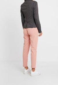 Person i grå tætsiddende blazer, lyserøde tapered bukser og hvide sneakers, der står mod en ensfarvet hvid baggrund med ryggen til.