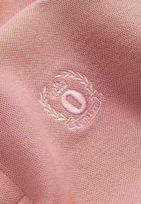 Gros plan sur un tissu rose texturé avec un emblème circulaire brodé en blanc comprenant une couronne et le mot "SPORT" en dessous.