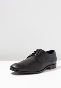 Zapato de vestir de cuero negro con un acabado texturizado, punta afilada y diseño de cordones. Presenta costuras laterales sutiles y un tacón bajo.
