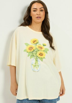 Kvinde iført løs, lysegul T-shirt med trykt klar vase, der holder lysegule solsikker og grønne blade.