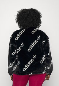 Černá plyšová flísová bunda s opakujícím se bílým logem "adidas", kulatým výstřihem a volným střihem. Hladká textura, dlouhé rukávy a spadlá ramena.