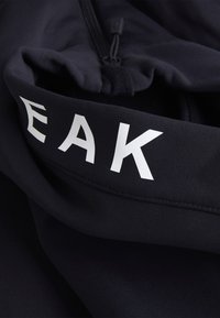 Sort stof med en glat tekstur, der pryder store hvide bogstaver, der staver "EAK." Inkluderer lynlås-lukning og elastiske detaljer.