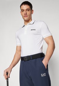 Biela polo košeľa s čiernymi akcentmi a logom "BOSS", v kombinácii s nohavicami námornej modrej farby s logom a čiernym opaskom. Jemne textúrovaná látka.