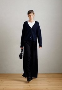 Ναυτικό πλεκτό ζακέτα με V-ανοιχτό λαιμό και τρία μαύρα κουμπιά, συνδυασμένη με μαύρες παντελόνες με φαρδιά πόδια και μια μικρή μαύρη τσάντα. Λείος υφή.