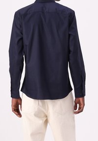 Navy blue long-sleeve shirt with a smooth texture, button cuffs, and a curved hem, worn with light-colored trousers.