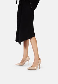 Tacchi alti in vernice beige con punta affilata e sottile tacco a stiletto, esposti accanto a un lungo cappotto nero.