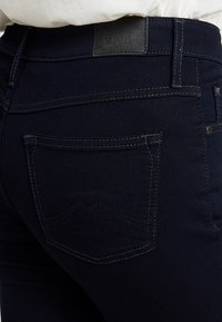Jeans de mezclilla azul oscuro con costuras visibles, dos bolsillos traseros y una etiqueta de marca de cuero en la cintura.
