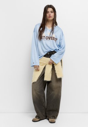 Lichtblauw langwerpig shirt met de tekst "GET OVER IT", oversized bruine baggy jeans en een crèmekleurige nepbont wikkel rond de taille. Beige schoenen.