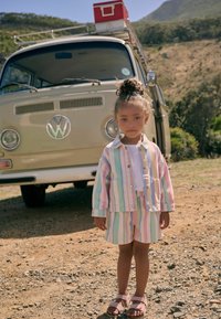 Flerfärgad randig outfit med en ljusblå jacka och shorts. Tyget är lättviktigt. Bruna sandaler kompletterar looken. En vintage Volkswagen-buss i bakgrunden.