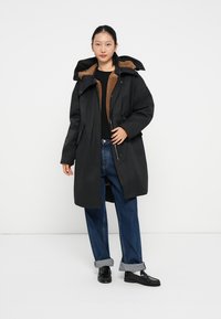Sort parka med en brun indvendig, der har hætte, frontlynlås og sidelommer. Båret over mørke jeans og sorte sko med opfoldet kant.