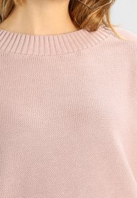 Mjukt rosa stickad tröja med ribbat rundhals, bärs av en person med ljust brunt hår synligt vid axeln.