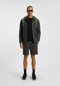 Felpa zip grigia scura con cappuccio e lacci, t-shirt nera, pantaloni corti abbinati e sneaker nere. Include occhiali da sole e dettagli con logo.