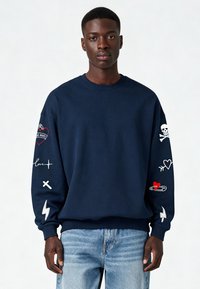 Sweatshirt bleu marine à manches longues avec divers motifs brodés, incluant des cœurs, un crâne et des éclairs. Tissu doux.