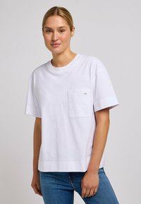 Camiseta de algodón blanca con cuello redondo, mangas cortas y un bolsillo en el pecho izquierdo que presenta una pequeña etiqueta de logo. Ajuste relajado.