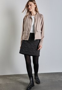 Helle beige Bomberjacke mit zwei Fronttaschen, kombiniert mit einem schwarzen Stepprock, schwarzen Strumpfhosen und Stiefeletten, vor einem neutralen Hintergrund.