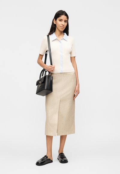 Γυναίκα που στέκεται, φοράει μπεζ φούστα, κοντομάνικο πουκάμισο με μπλε γιακά, μαύρα μοκασίνια και κρατάει μια μαύρη τσάντα ώμου.