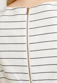 Close-up van een wit kledingstuk met dunne zwarte horizontale strepen en een centrale gouden rits op de achterkant nabij de nek.