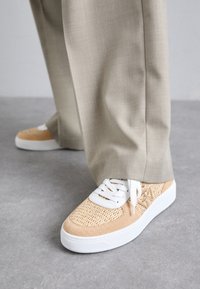 Beige Sneaker mit einem strukturierten gewebten Obermaterial und Wildlederakzenten, weißen Schnürsenkeln und Sohle, kombiniert mit hellgrauen Hosen.