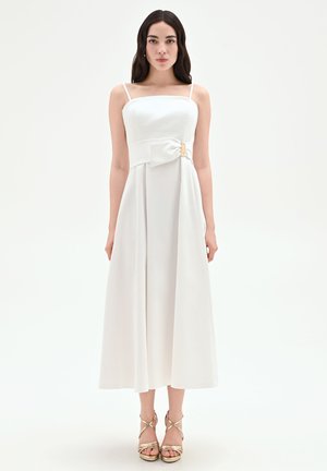 Femme portant une robe midi blanche sans manches avec un noeud à la taille et des sandales dorées à talons hauts à brides, debout.