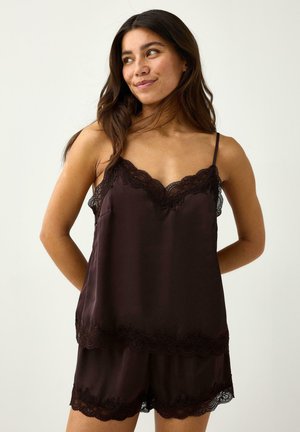 Kvinde med langt mørkt hår iført mørkebrun silkecamisole og matchende shorts med blonder, kigger til siden mod en ensfarvet baggrund.