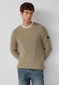 Beige geripptes Sweatshirt aus strukturiertem Stoff, mit einem runden Ausschnitt und einem kontrastierenden weißen Innenkragen, sowie einem kleinen Logo auf dem Ärmel.
