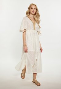 Witte kanten jurk met franjes, V-halslijn en gouden details. Lange mouwen en een vloeiende silhouette, gecombineerd met metallic sandalen.
