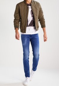 Olivově zelená prošívaná bomber bunda, bílý tričko s grafikou, modré taperované džíny a bílé tenisky. Hladká textura a přiléhavý design.