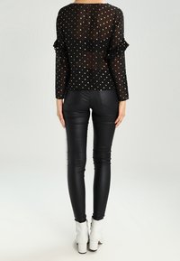 Blusa nera trasparente con pois dorati e maniche a volant, abbinata a pantaloni neri aderenti e lucidi e stivaletti bianchi alla caviglia.