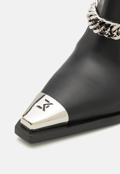Black leather ankle boot featuring a pointed toe with a metallic silver cap and a decorative silver chain accent around the ankle.