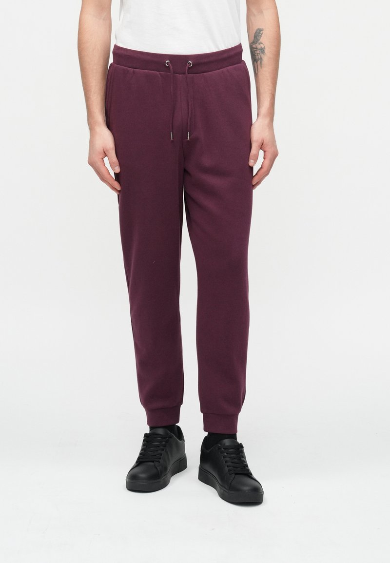 Μπορντό φόρμα με ελαστική μέση με κορδόνι, ριμπ μανσέτες στους αστραγάλους και πλαϊνές τσέπες, φορεμένη με μαύρα αθλητικά παπούτσια σε ουδέτερο φόντο.