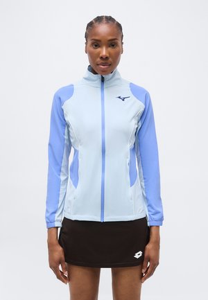 Femeie în picioare, cu fața spre înainte, purtând o jachetă cu fermoar de culoare albastru deschis și lavandă și o fustă sport neagră, pe un fundal simplu.