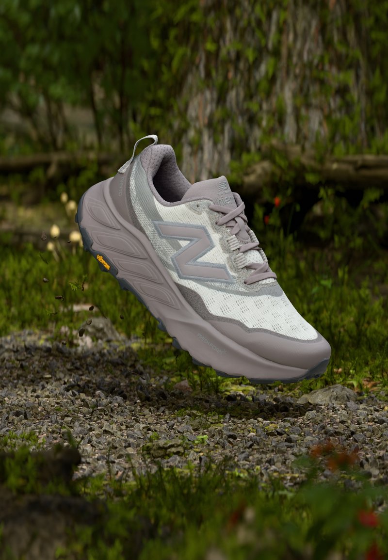 Hellgrauer Wanderschuh mit Mesh-Obermaterial, violetten Akzenten, gepolsterter Sohle und Vibram-Außensohle, schwebt über einem felsigen, grünen Gelände.