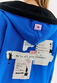 Niebieska bluza z kapturem z czarnymi akcentami, z grafikami w stylu kolażu, w tym tekstem i ilustracjami na plecach. Tekst brzmi: "Wszyscy gdzieś zmierzamy."