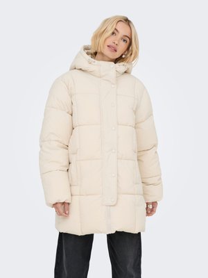 Femme blonde portant un manteau matelassé beige à capuche avec des boutons sur le devant, debout devant un fond blanc uni.