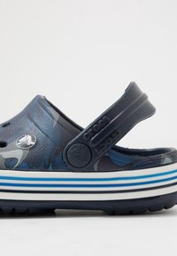 Modré maskáčové dřeváky se zadním páskem, vyrobené z gumy. Mají perforace a podrážku s bílými a modrými pruhy. Viditelný detail s logem.