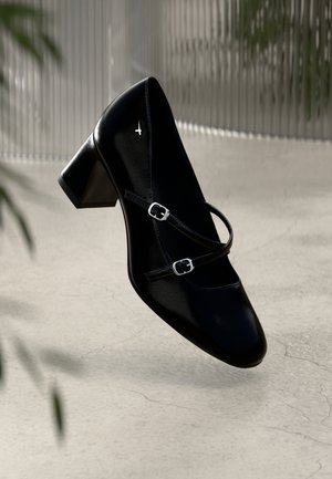 Chaussure à talon en cuir noir avec un bout carré, deux sangles décoratives et des boucles en argent. Texture lisse avec une finition brillante.