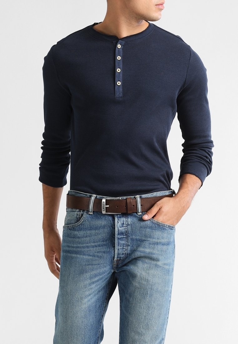 Homme en chemise henley bleu marine à manches longues avec quatre boutons, jean bleu et ceinture marron, main dans la poche, sur un fond uni.